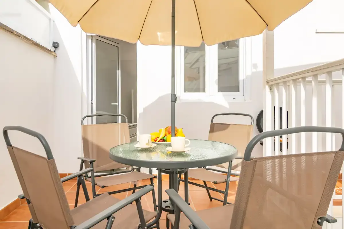 Un pequeño patio exterior tiene una mesa redonda y asientos bajo una sombrilla grande.