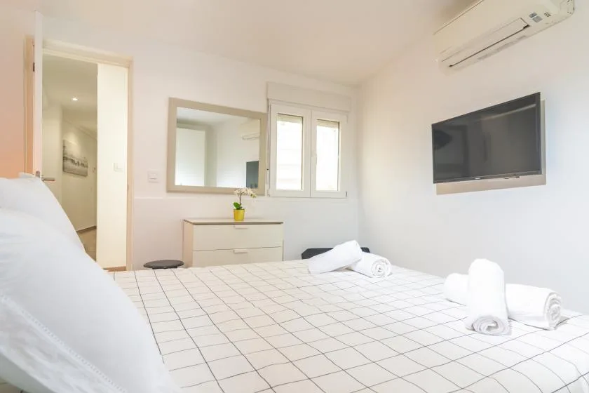 Un dormitorio luminoso con una cama bien tendida y un televisor en la pared.