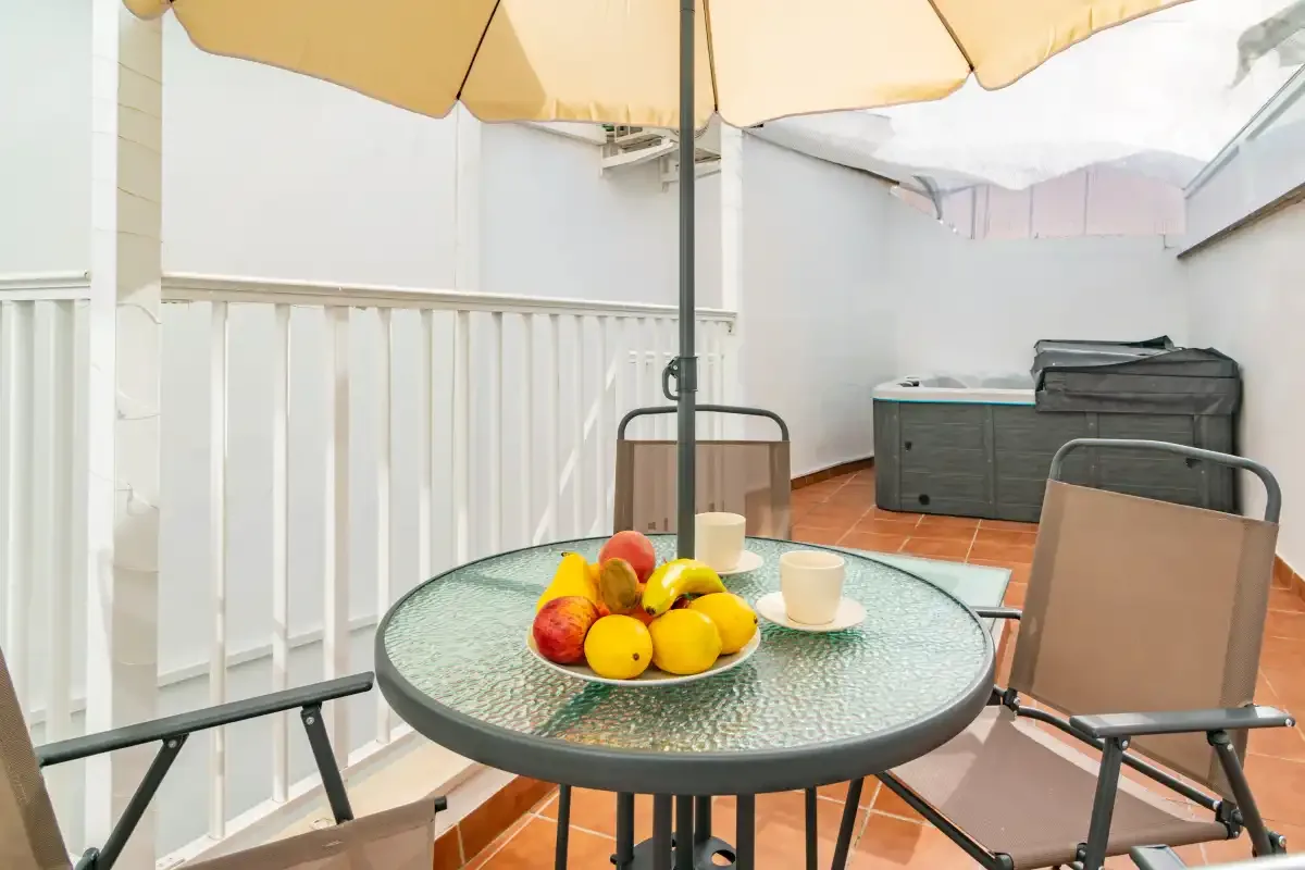 Una zona de comedor en el patio con mesa de vidrio, asientos y una sombrilla.