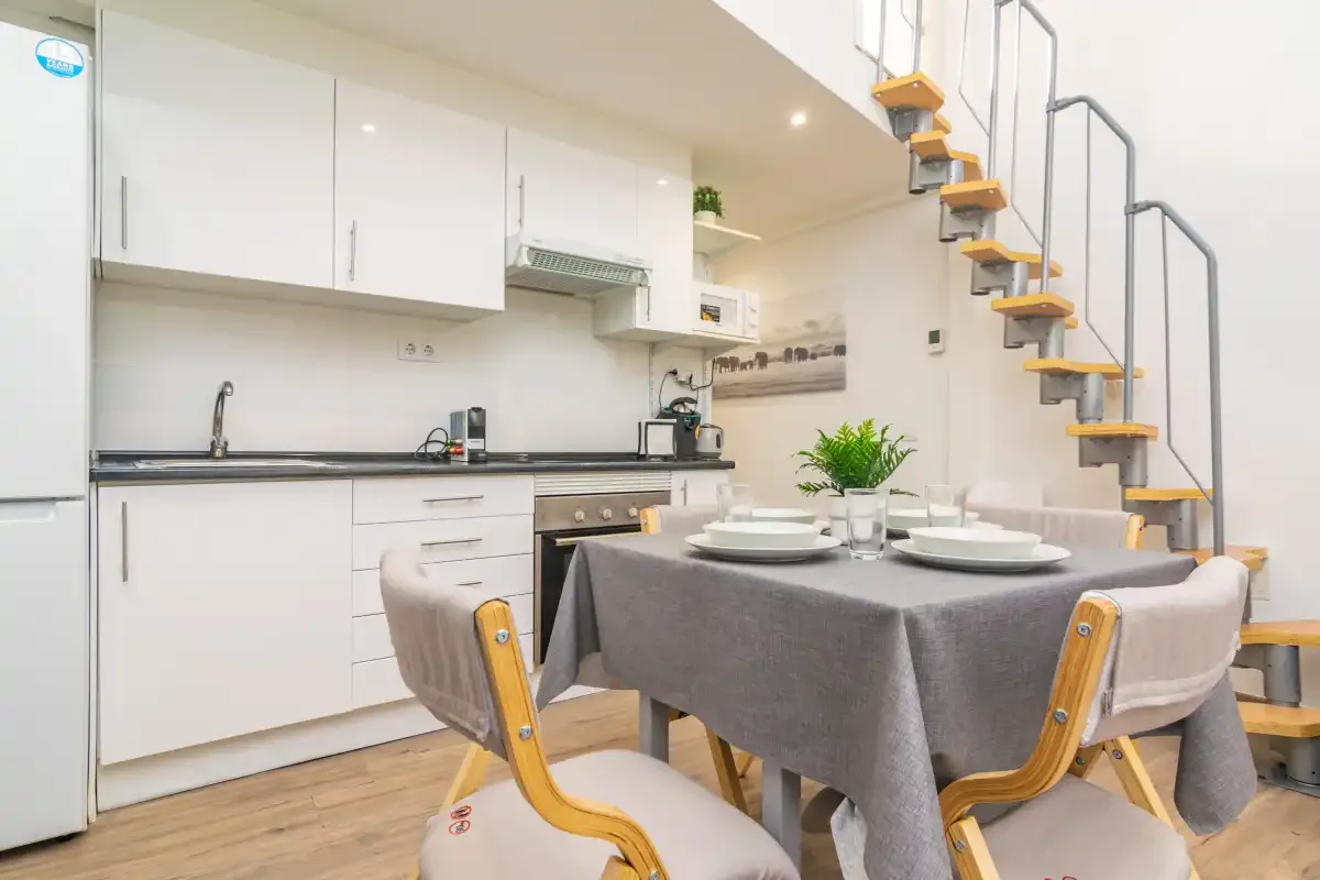 Una cocina luminosa con zona de comedor y una mesa bajo una escalera abierta.