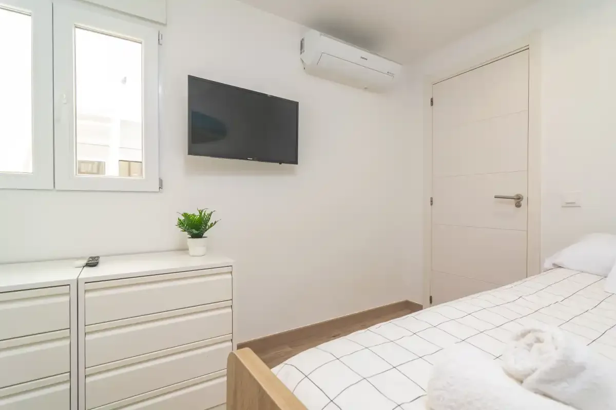 Un dormitorio luminoso con una cama, una cómoda y un televisor en la pared.