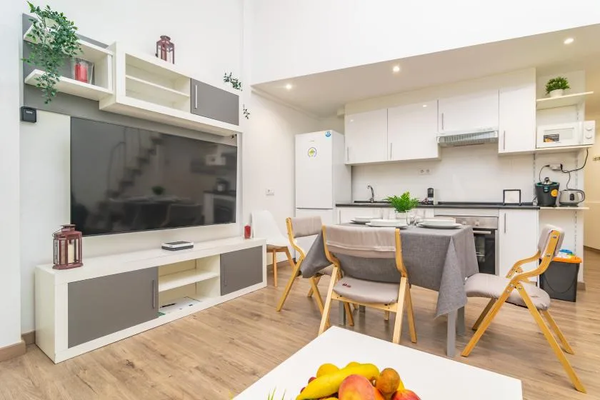 Una luminosa sala-comedor comunica con una cocina blanca, con mesa y asientos.