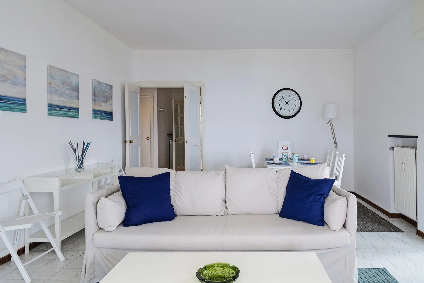 Una sala de estar luminosa con un sofá, una mesa blanca y cuadros enmarcados.