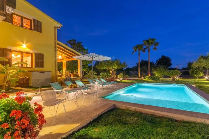 An illuminated outdoor swimming pool beside a house and patio seating at dusk.