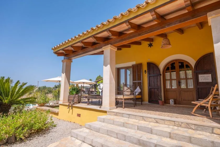 A sunny yellow house porch with wooden doors and a shaded seating area.