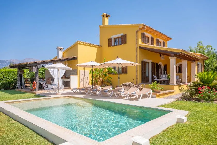 A yellow house overlooks a swimming pool, with patio seating beside the water.
