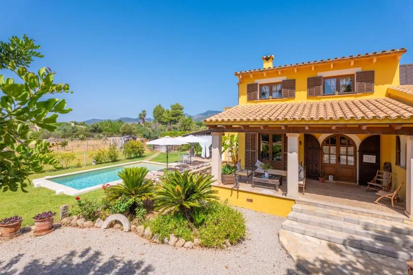 A yellow house with a covered patio overlooks a swimming pool and landscaped garden.