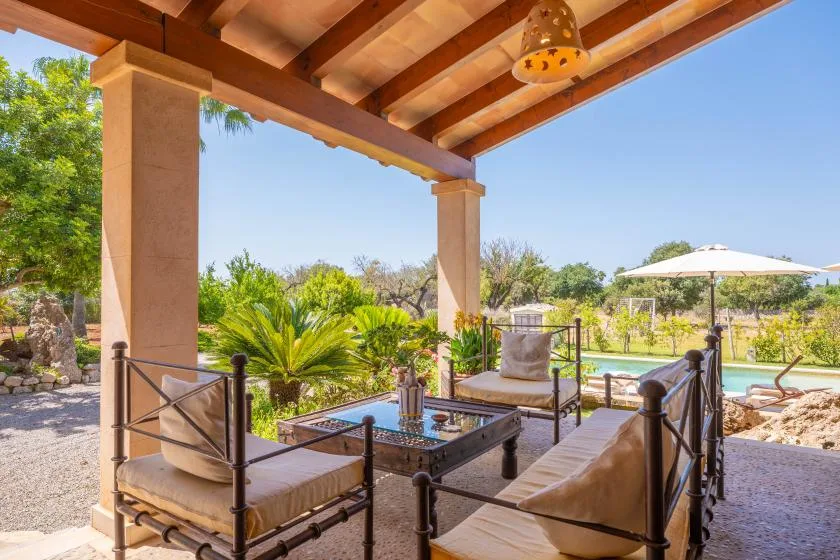 A covered patio with cushioned seating and a table overlooks a garden and pool.