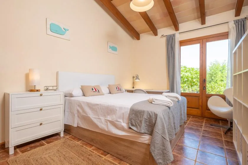 A bright bedroom with a large bed, bedside furniture, and wooden ceiling beams.