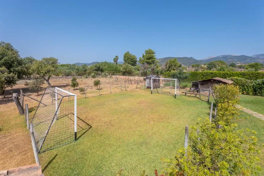 An outdoor grassy field with soccer goals, trees, and distant hills.