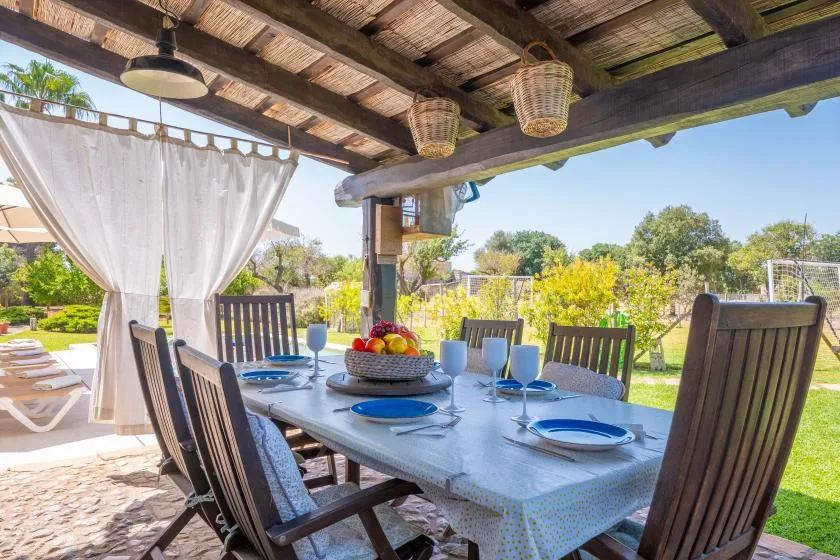 An outdoor dining area sits beneath a covered pergola, with a set table overlooking greenery.