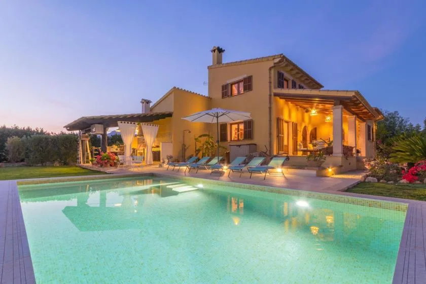 A lit villa stands beside a swimming pool, with outdoor seating on the patio.