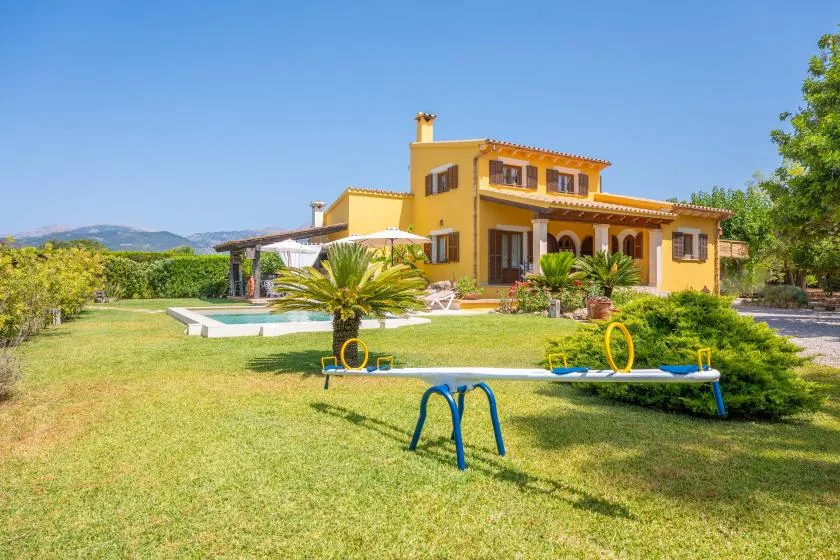A yellow house stands behind a grassy yard with a swimming pool and outdoor play equipment.