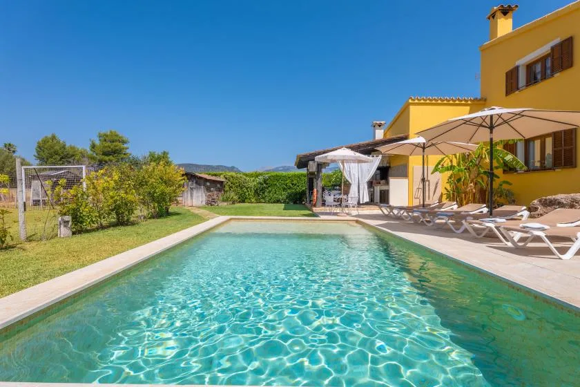 A swimming pool beside a yellow building, with lounge seating and umbrellas along the deck.