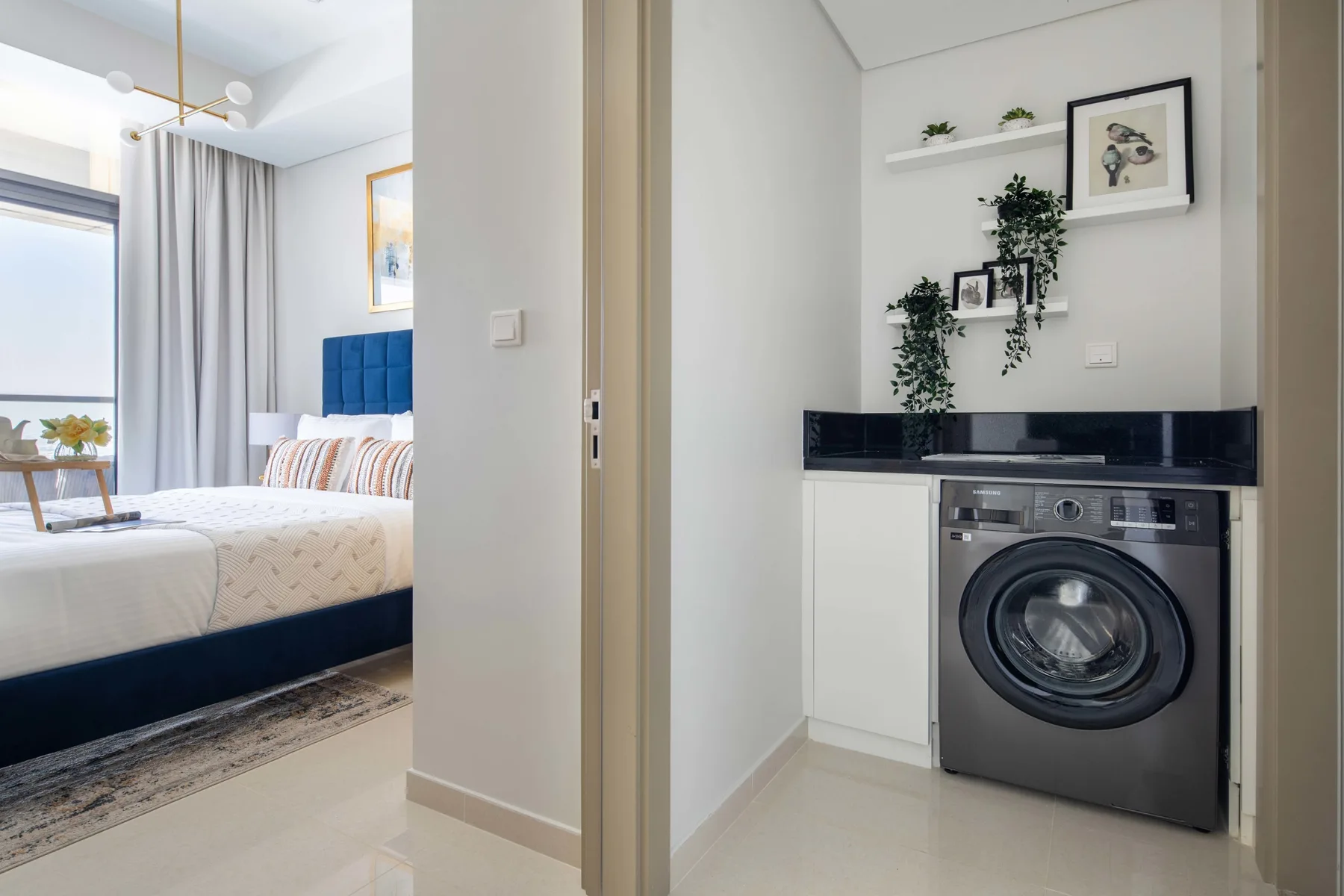 A bedroom with a neatly made bed sits beside a laundry area with a front-loading washing machine.