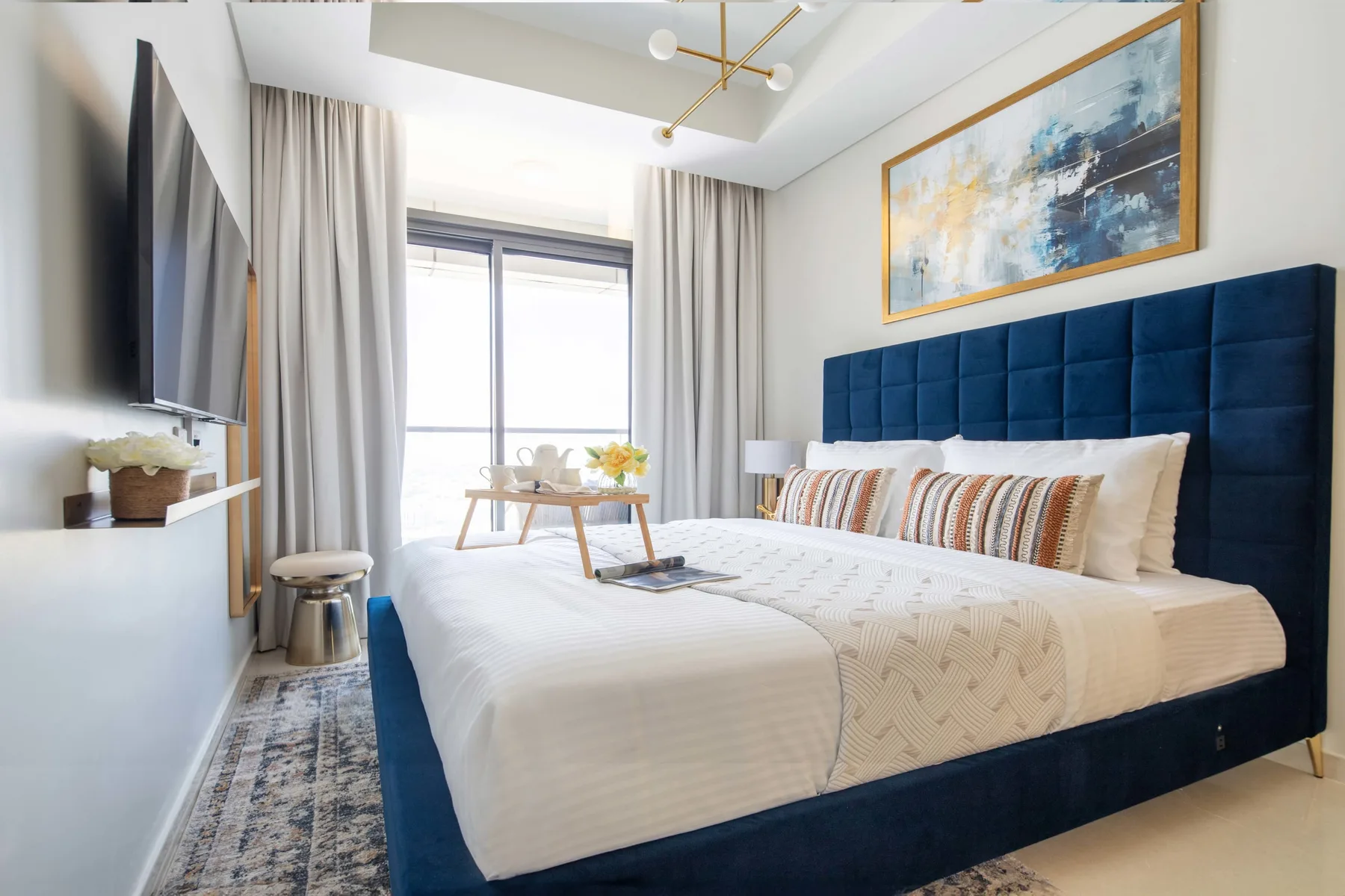 A bright bedroom with a large bed, blue upholstered headboard, and window.
