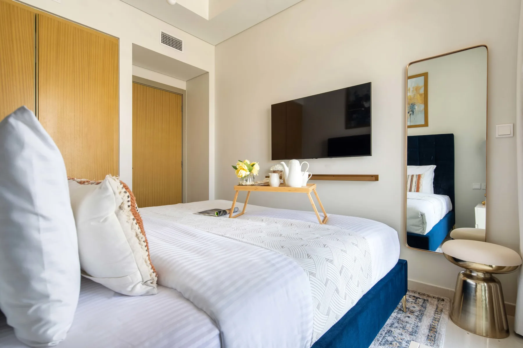A bedroom with a neatly made bed, a wall-mounted television, and a full-length mirror.