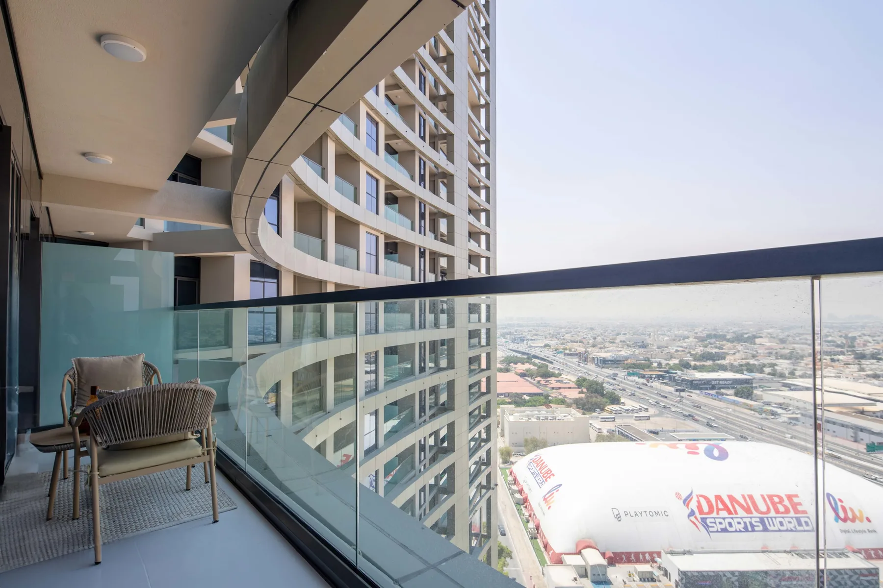 A high-rise balcony overlooks a cityscape, with glass railings and outdoor seating.