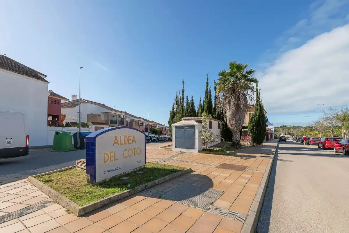 Entrada de una calle residencial con un letrero, aceras, coches estacionados y palmeras.