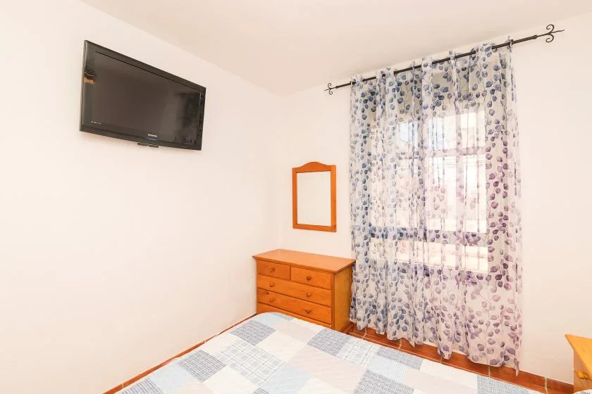 Un dormitorio con cama, cómoda, televisor de pared y ventana con cortinas.