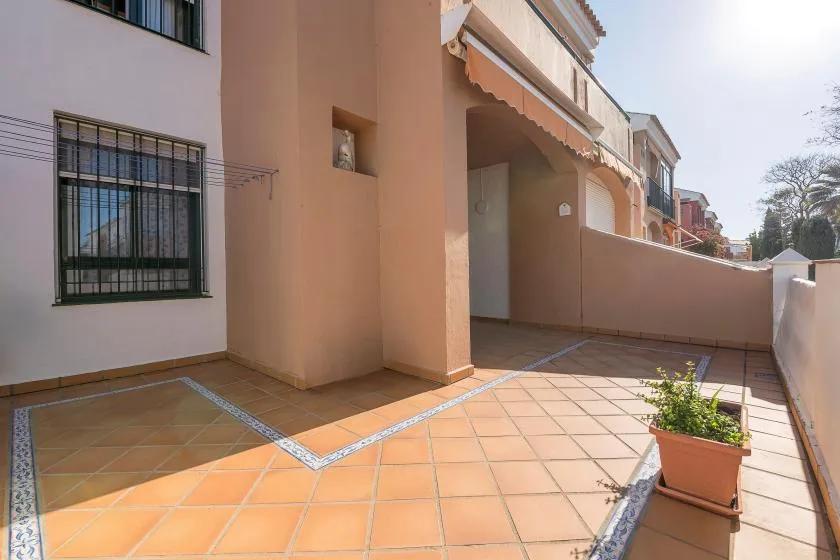 Una terraza soleada con baldosas junto a un edificio color durazno y una planta en maceta.