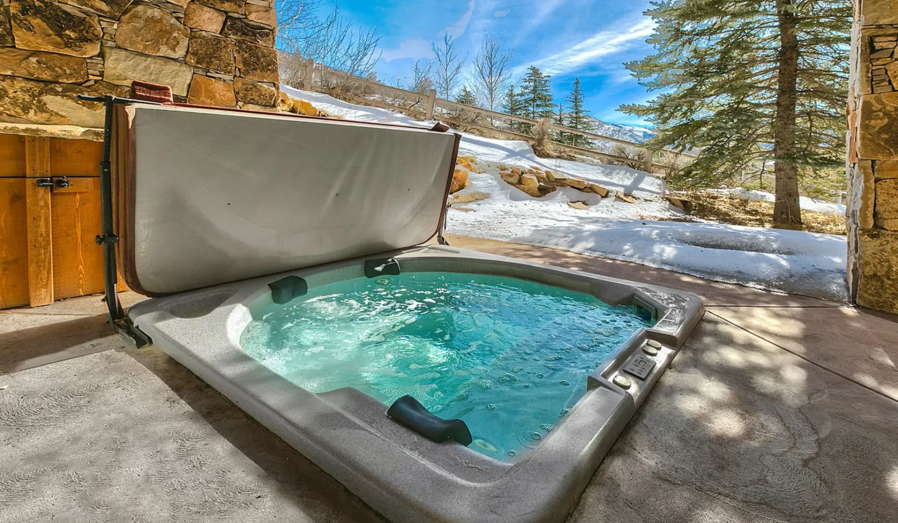 Una bañera de hidromasaje al aire libre está en un patio junto a un muro de piedra, con nieve al fondo.