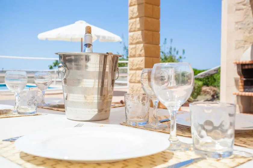 A table is set with glassware and a metal ice bucket beside a sunny pool.