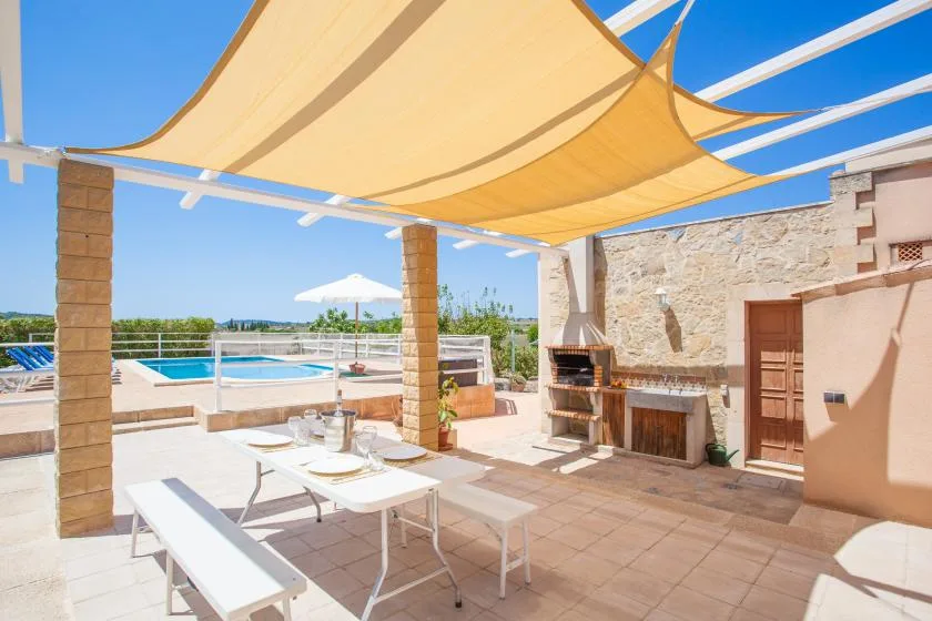 A sunny outdoor patio has dining seating, a built-in grill, and a swimming pool.