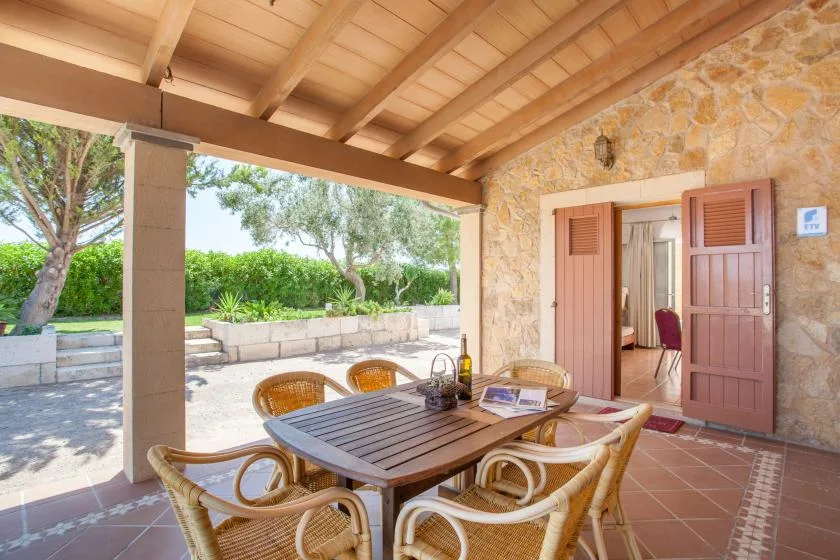 A covered stone patio has a wooden dining table and seating beside open doors.