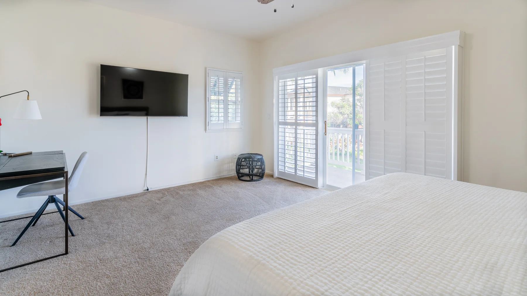 A bright bedroom with a bed, a wall-mounted television, and windows with shutters.