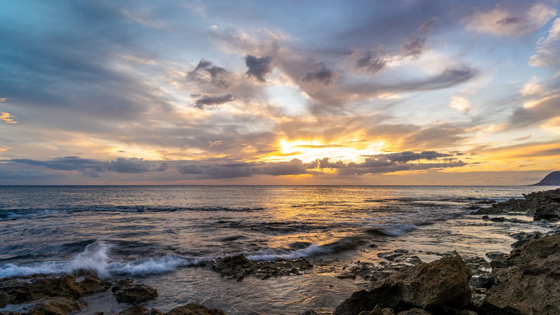 A rocky shoreline and ocean waves sit beneath a colorful sunset sky.