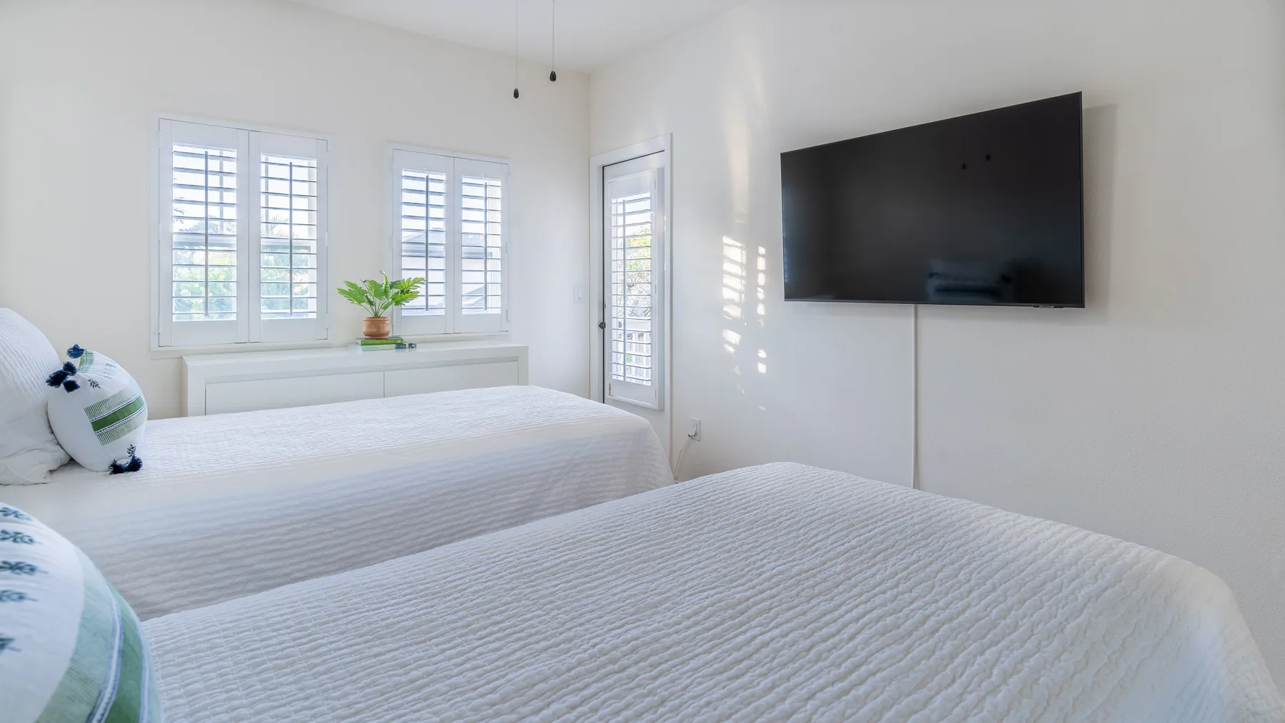 A bright bedroom with two beds, shuttered windows, and a wall-mounted television.