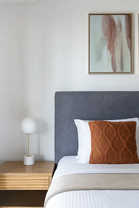 Un dormitorio con una cama tendida, un cojín color óxido con textura y una lámpara de noche.