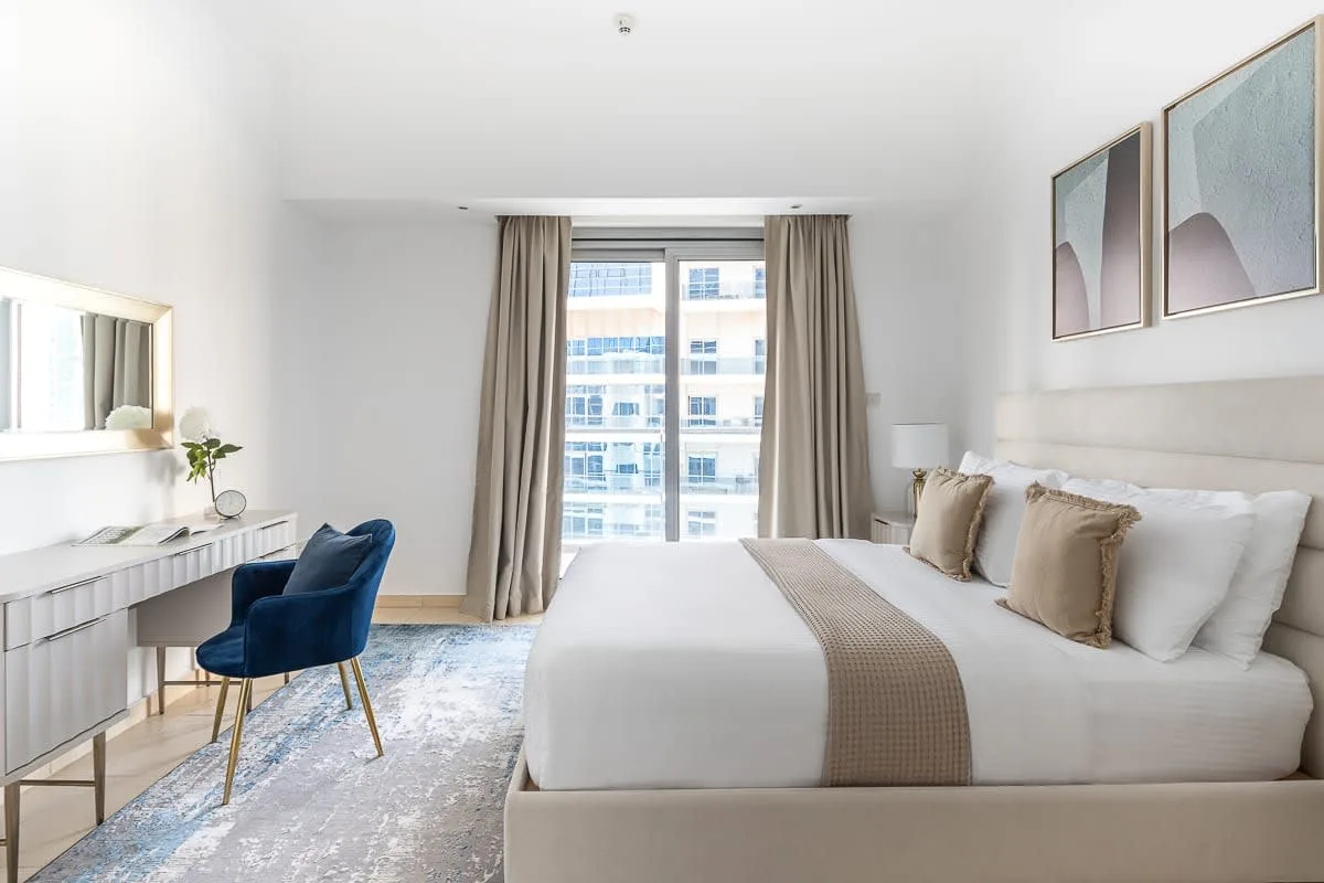 Dormitorio luminoso con una cama grande, un escritorio y una ventana con cortinas.