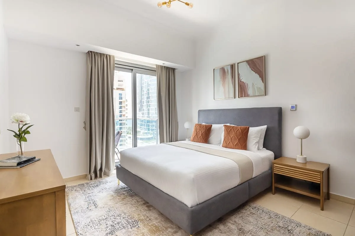 Un dormitorio luminoso tiene una cama bien hecha junto a una ventana grande.