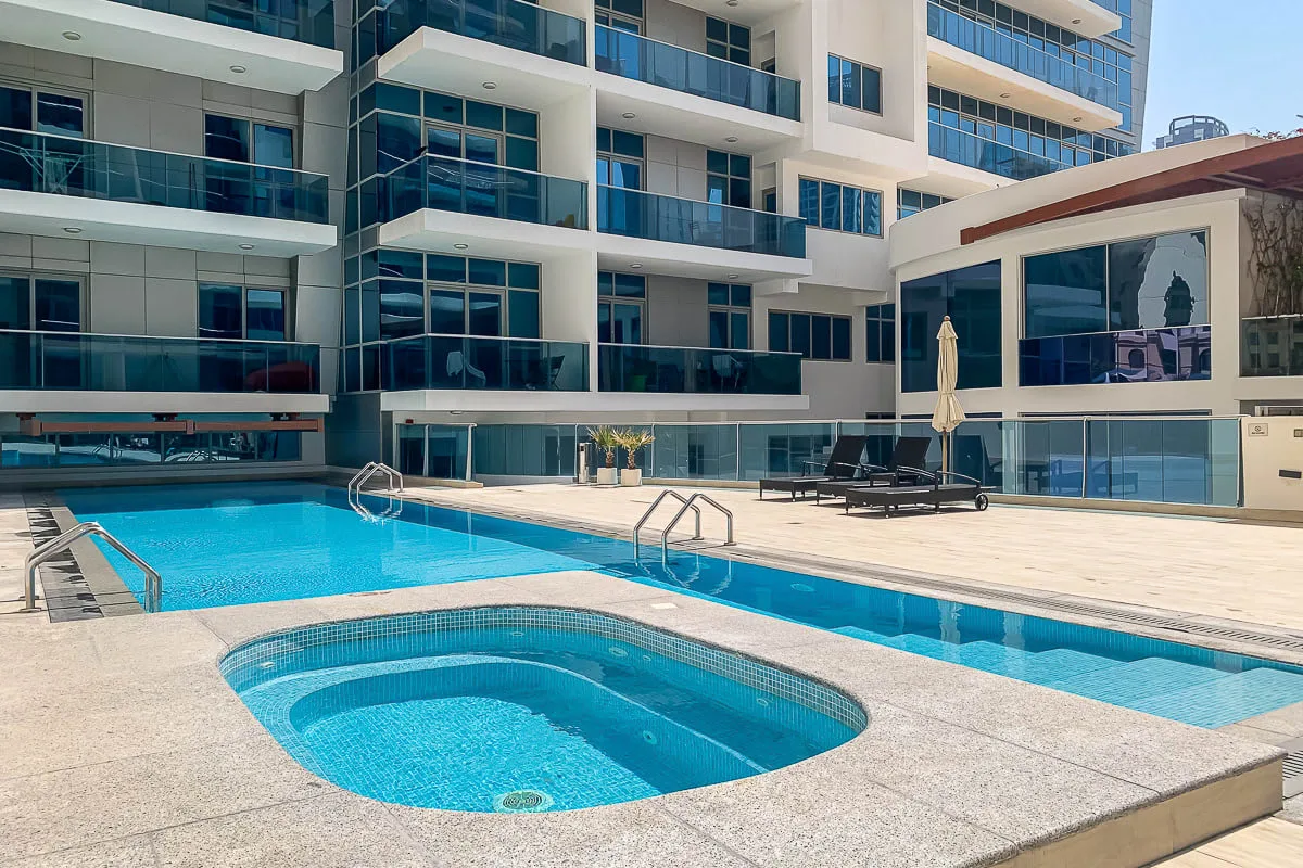 Una piscina al aire libre y otra más pequeña están junto a un edificio moderno de varios pisos.