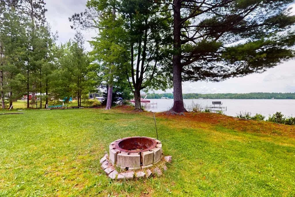 Un patio junto al lago con césped y un fogón circular bajo árboles altos.