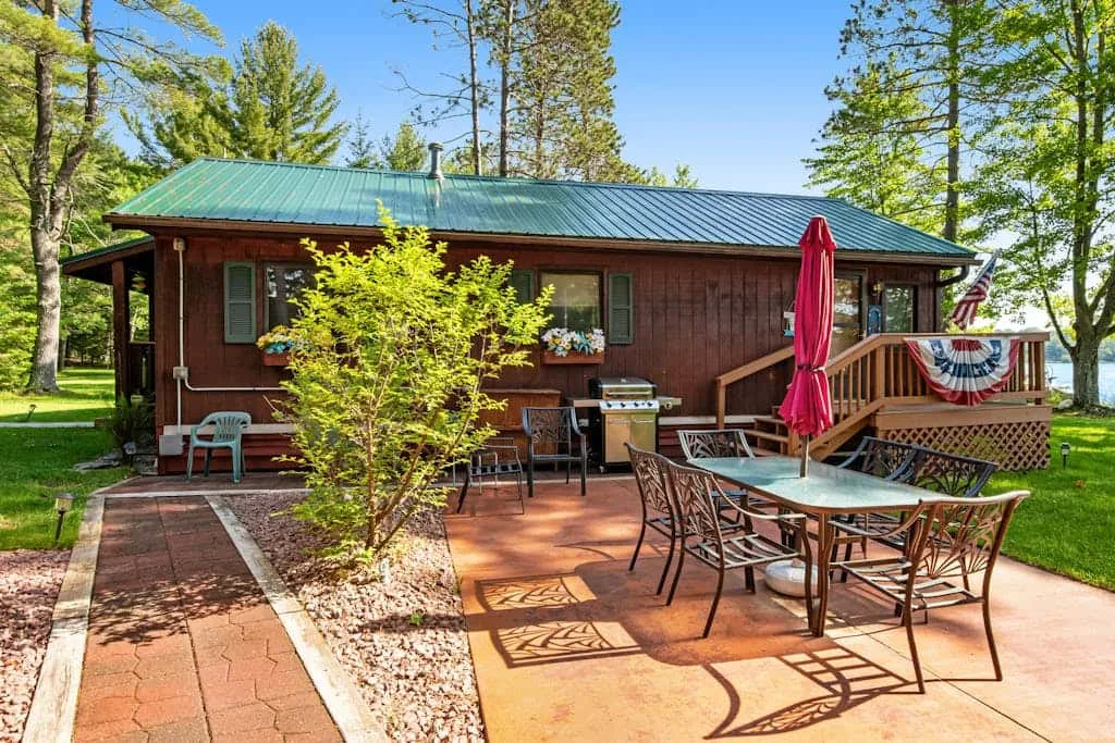 Una cabaña con patio pavimentado, mesa de comedor al aire libre y terraza rodeada de árboles.
