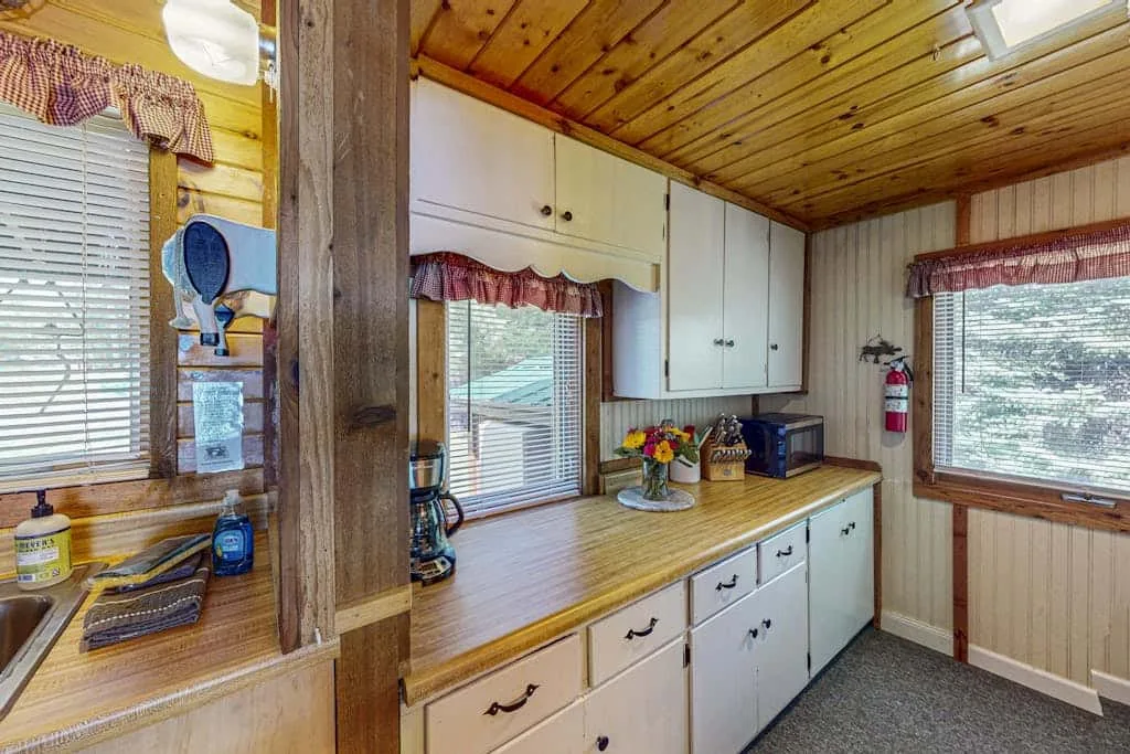 Una cocina revestida de madera con gabinetes blancos y una encimera larga bajo las ventanas.