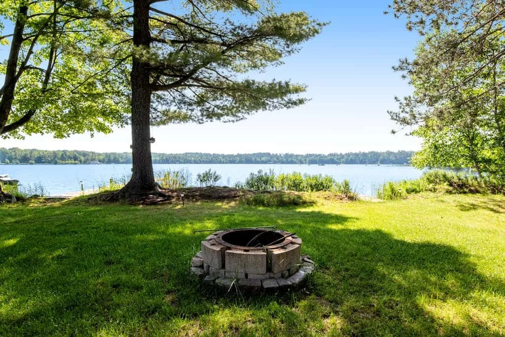Un patio junto al lago, con césped, árboles frondosos y un fogón circular.
