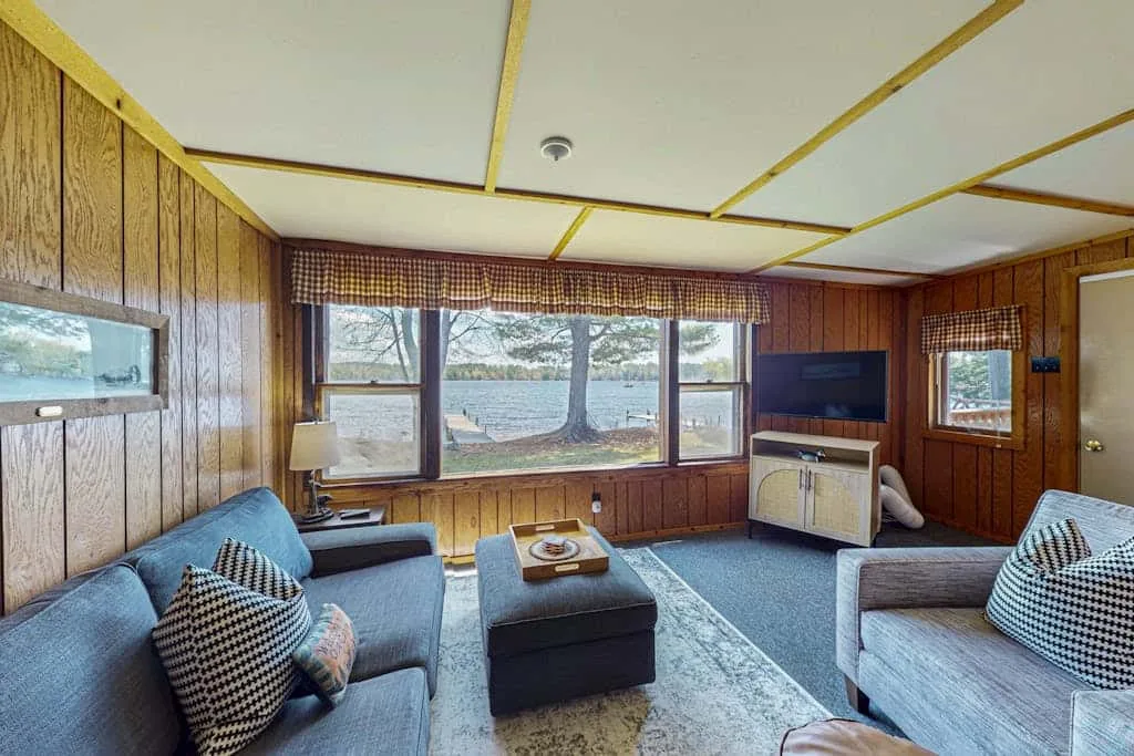 A wood-paneled living room with seating, a television, and windows overlooking the water.