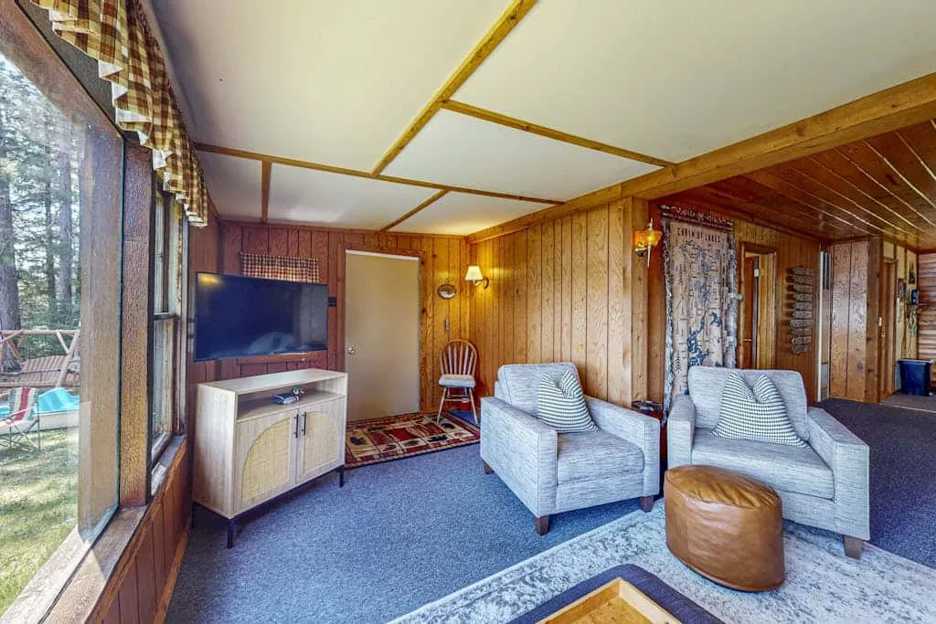 Sala de estar revestida de madera, con dos sillones, un televisor y grandes ventanas.