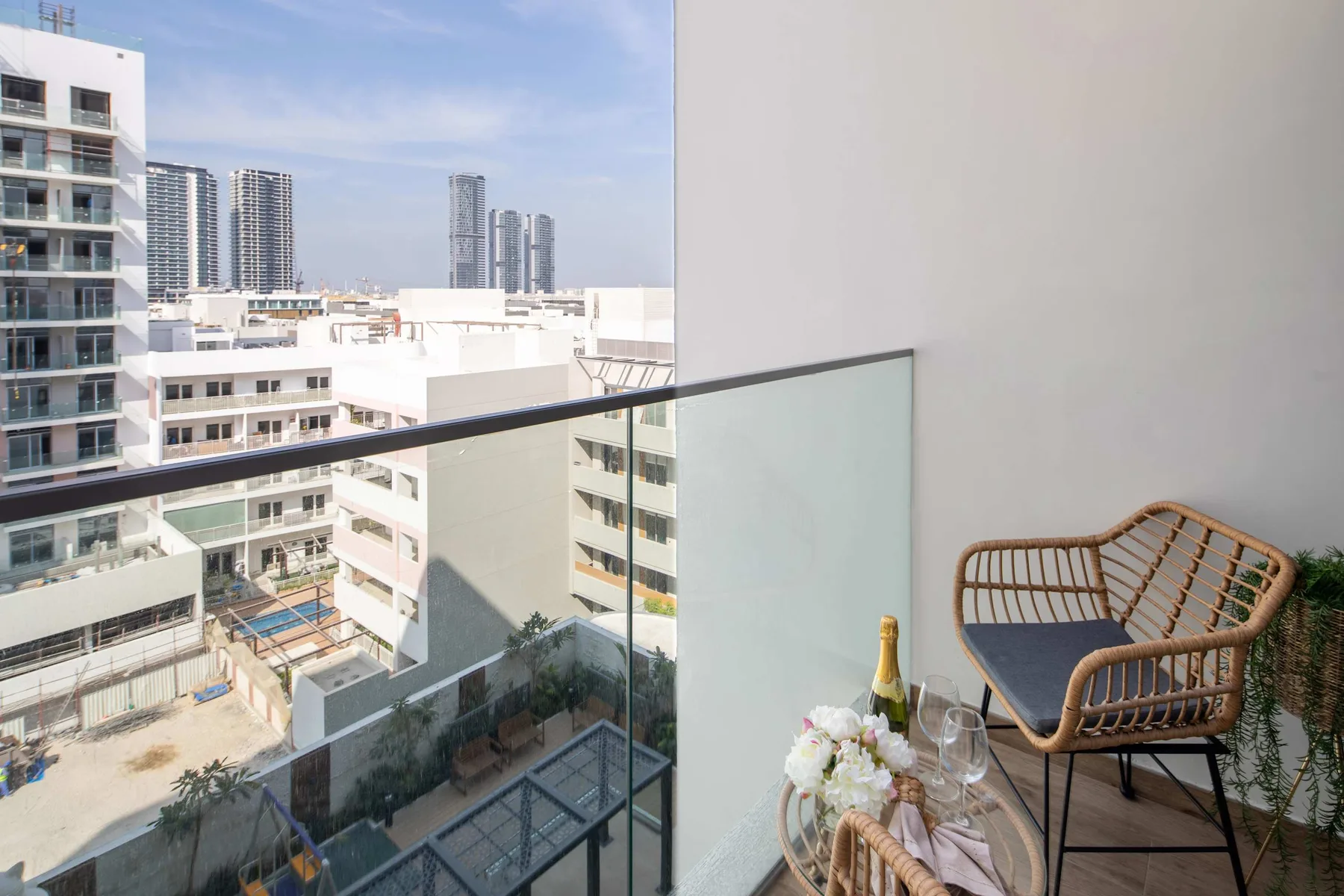 Un balcón da a edificios modernos, con una silla de mimbre y una mesa junto a la barandilla de cristal.