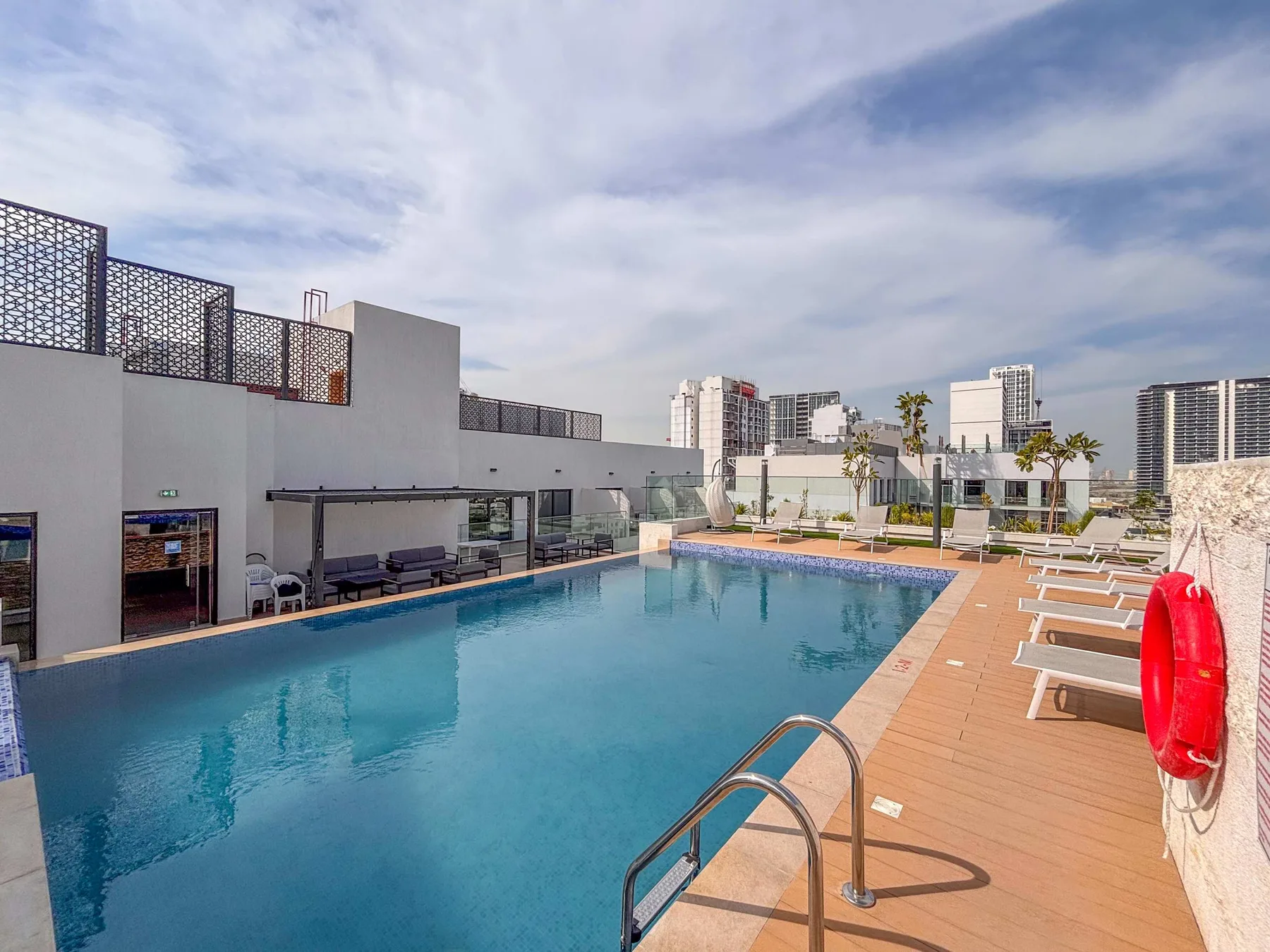 Una piscina en la azotea junto a una terraza de madera, tumbonas y edificios de la ciudad.
