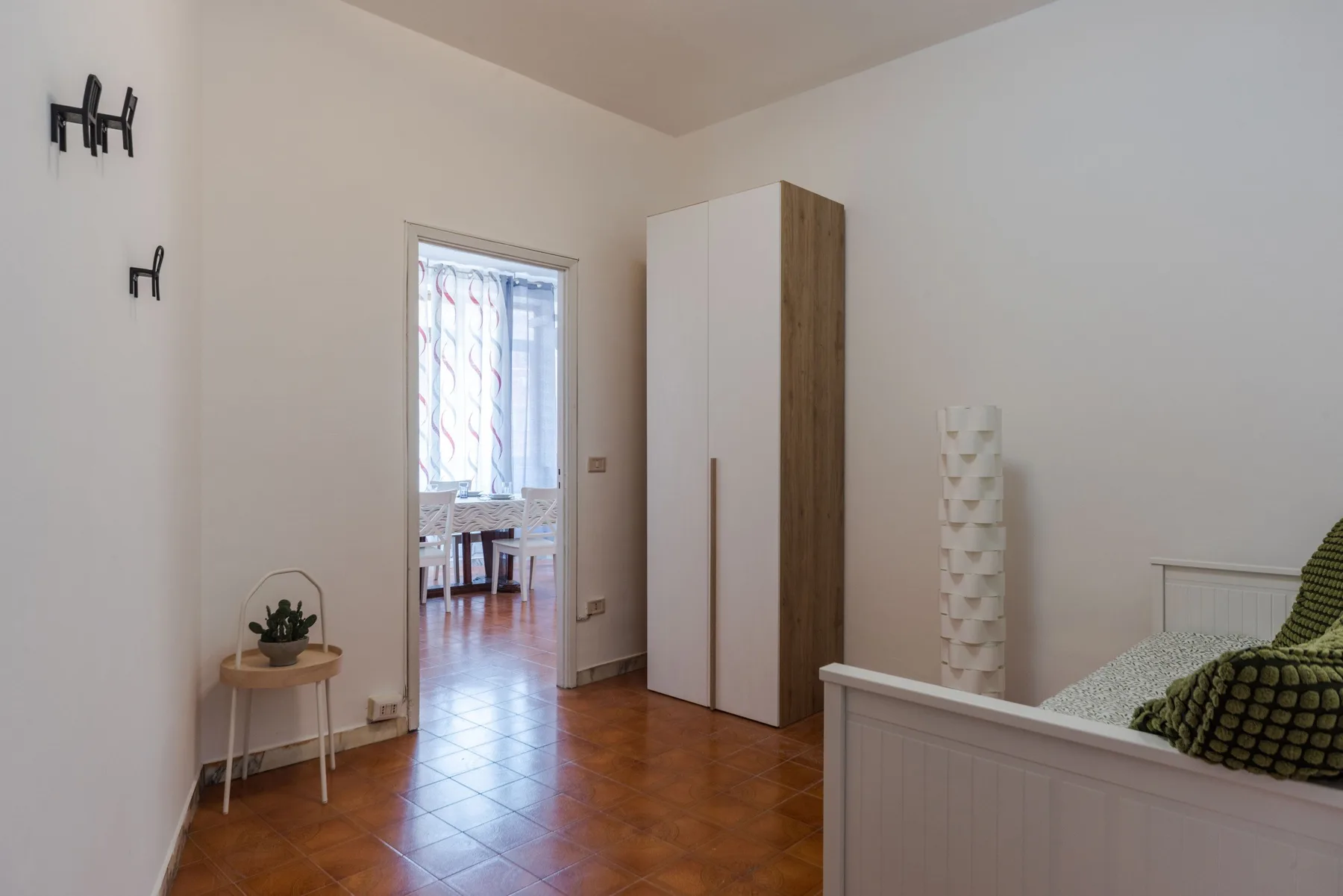 A bright bedroom with a tall wardrobe and an open doorway to a dining area.