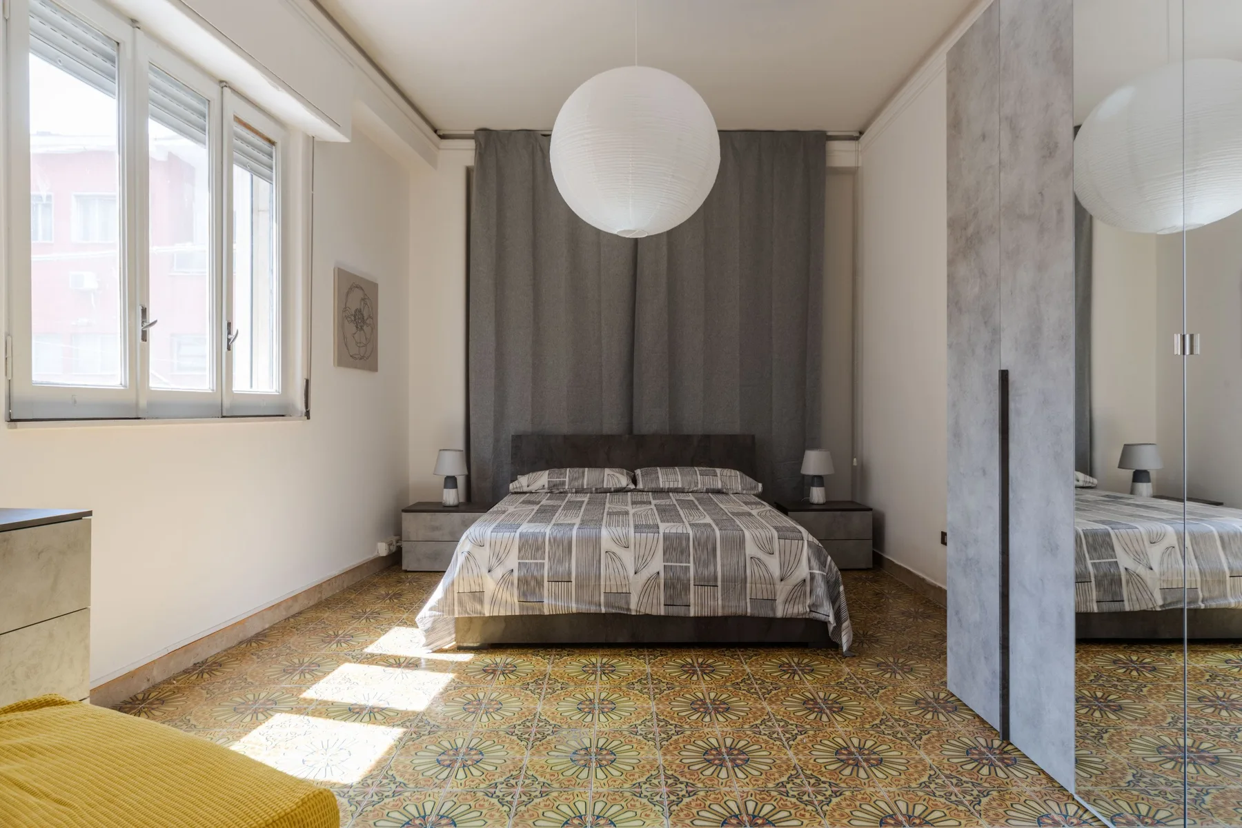 A bedroom with a bed centered in front of gray curtains and a window to the side.