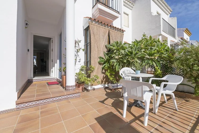 A sunny tiled patio with white outdoor seating and leafy plants beside the house.