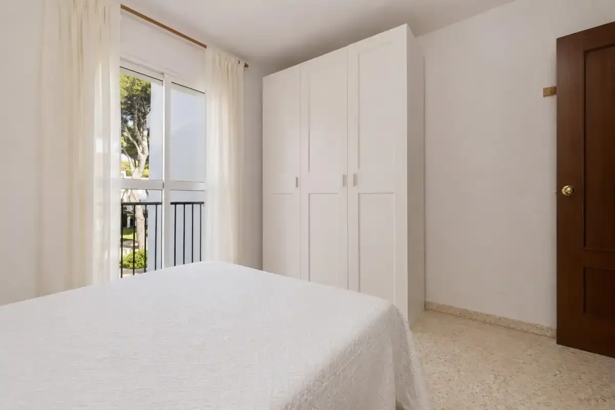 A bright bedroom with a bed, white wardrobe, and window opening onto a balcony.