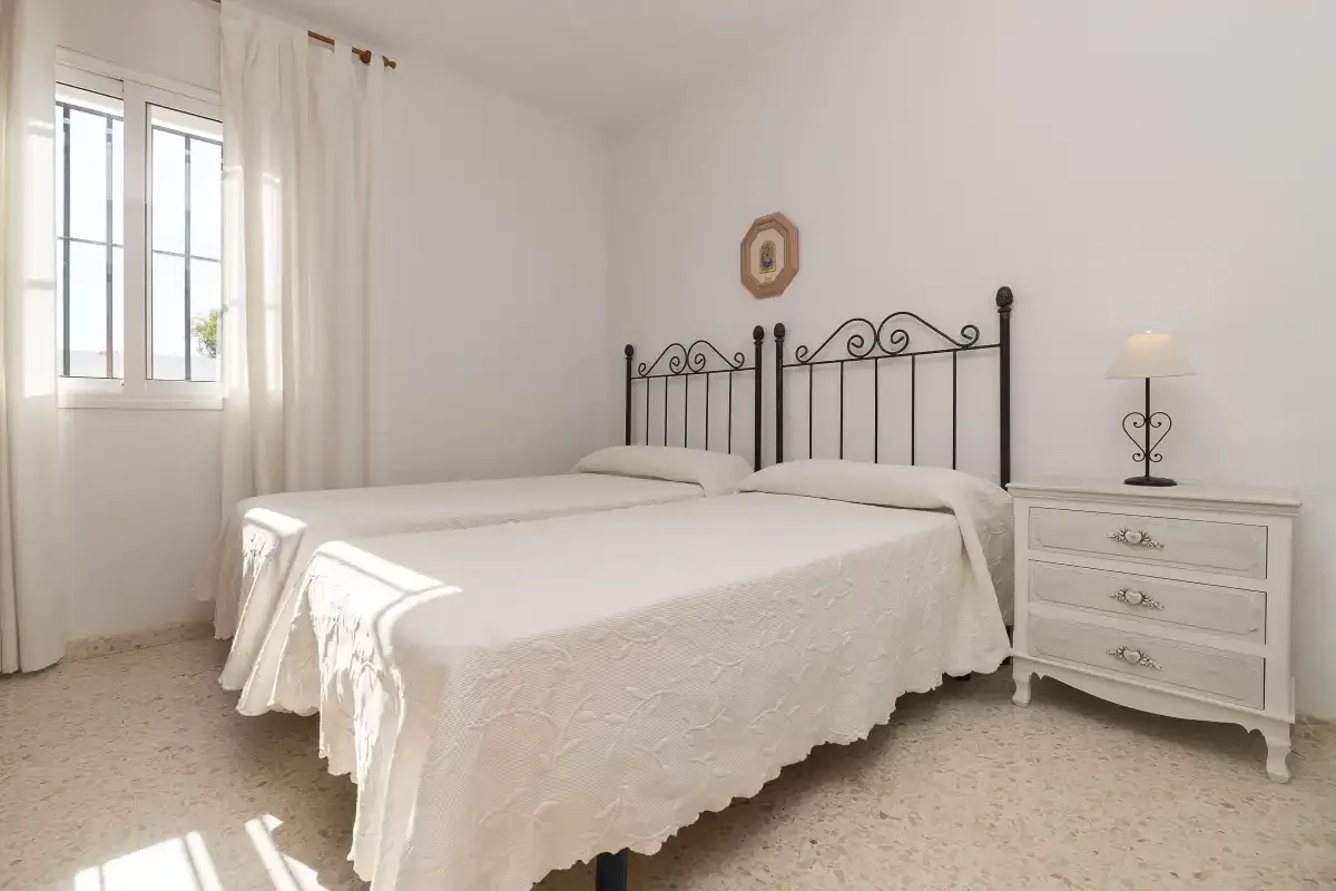 A bright bedroom with a double bed, bedside dresser, and window.