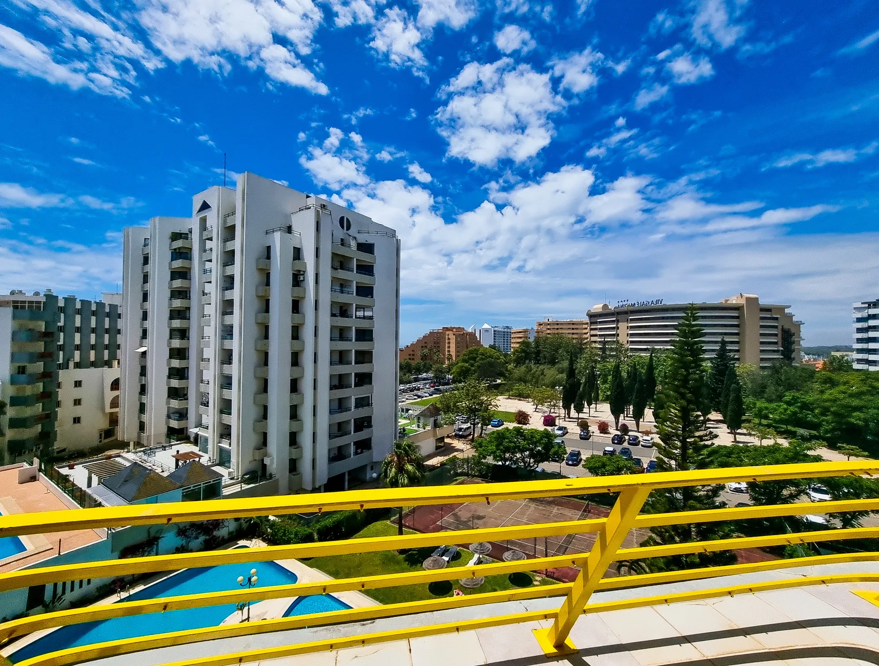 Un balcón da a edificios de apartamentos cercanos, árboles y una piscina.
